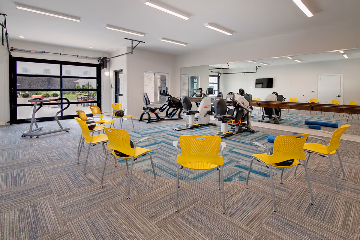 A bright exercise room with yellow chairs arranged in a circle on a carpeted floor. The room features exercise equipment including stationary bikes and a treadmill. Large windows and a glass garage-style door allow natural light to enter. A large mirror covers one wall, reflecting the room and equipment.