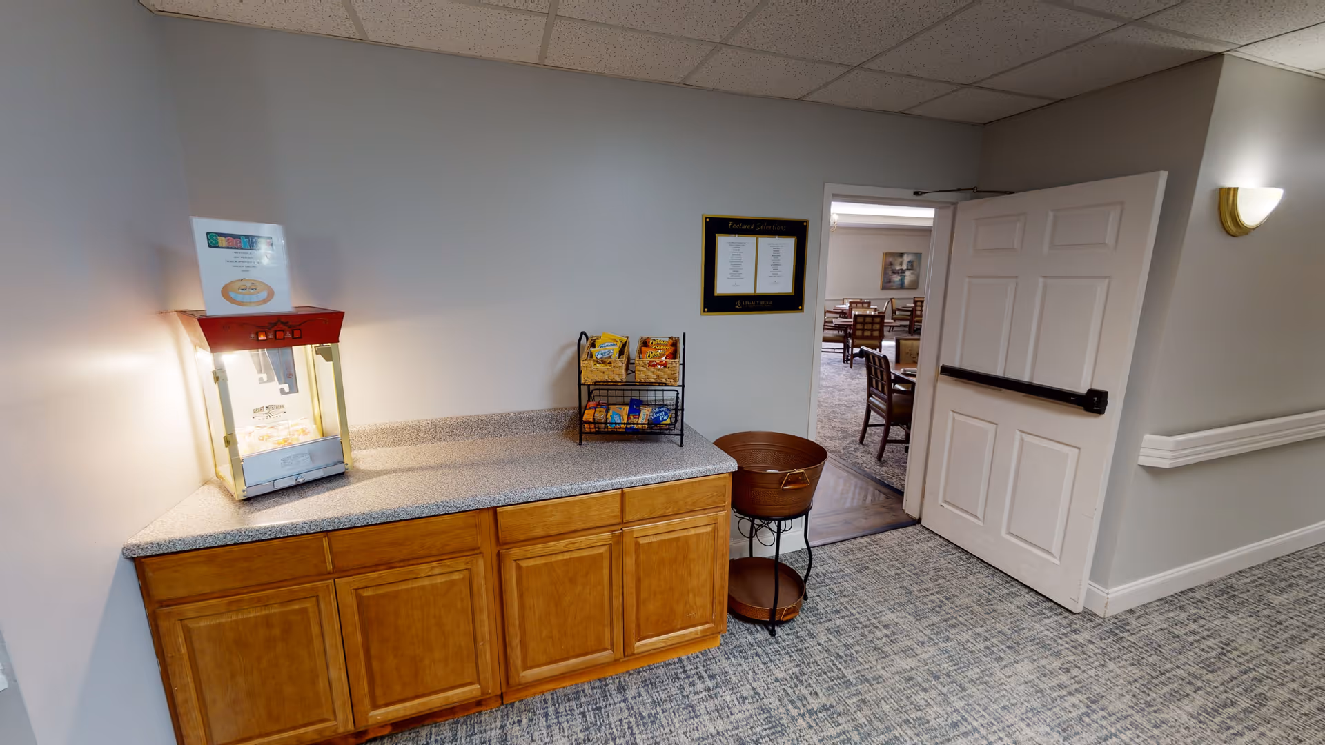 A small snack station in a senior living facility hallway with a popcorn machine on a countertop, a rack holding various snack bags, and a two-tiered round metal stand. An open door leads to a dining area with tables and chairs. The walls are light gray with a wall sconce light and a framed menu or notice on the wall.