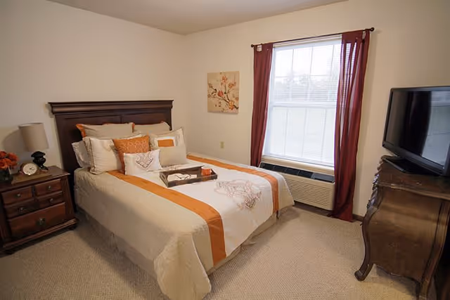 A neatly made bed with white and orange bedding in a bedroom. The bed has multiple pillows and a tray with a book and a cup on it. There is a wooden nightstand with a lamp, clock, and flowers on the left side of the bed. A window with red curtains and blinds is on the right wall, and a wooden dresser with a TV on top is positioned near the window. The room has beige carpet and light-colored walls with a floral painting hanging above the bed.