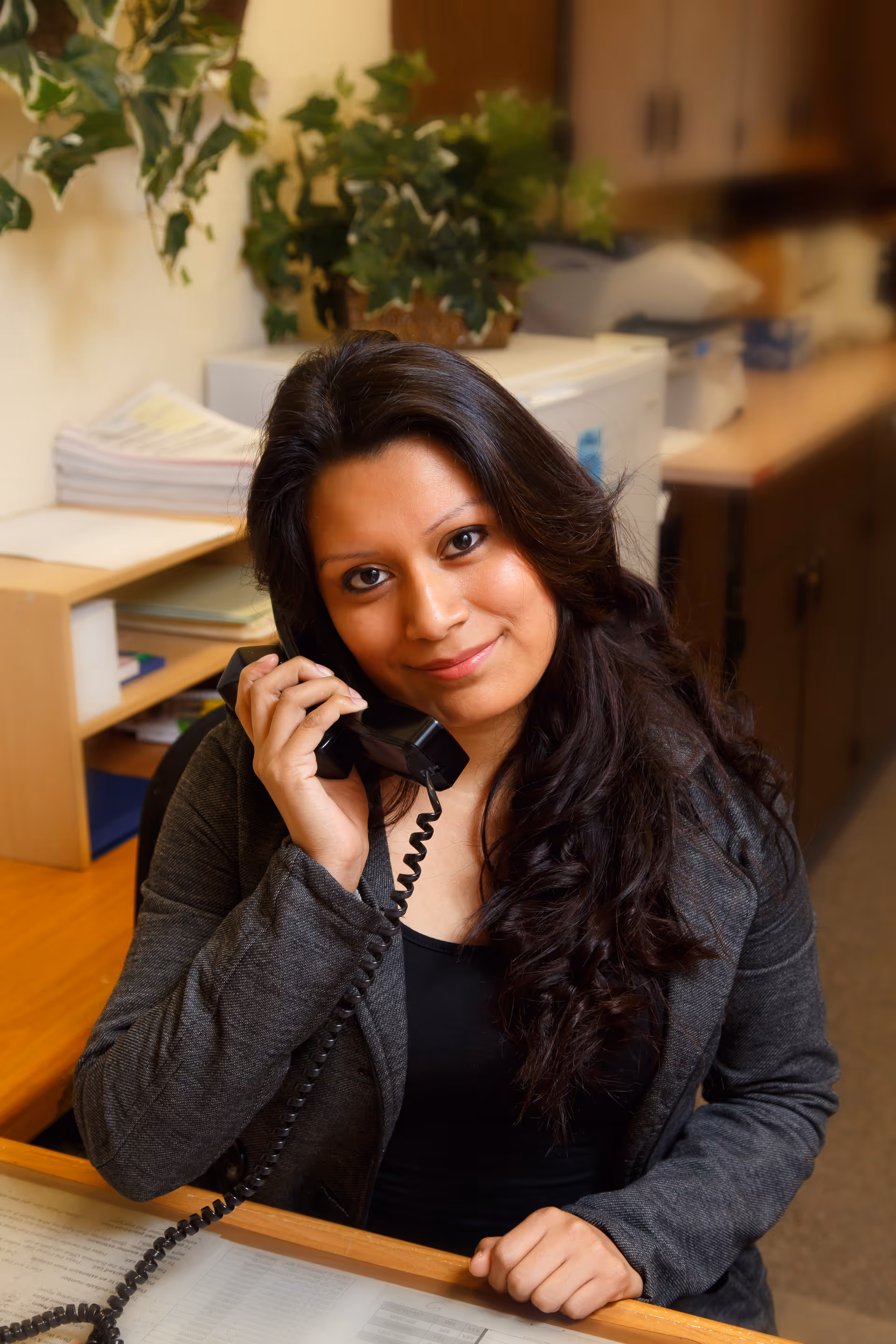 A woman with long dark hair sitting at a desk, smiling and holding a telephone receiver to her ear in an office setting with shelves, papers, and a printer in the background.
