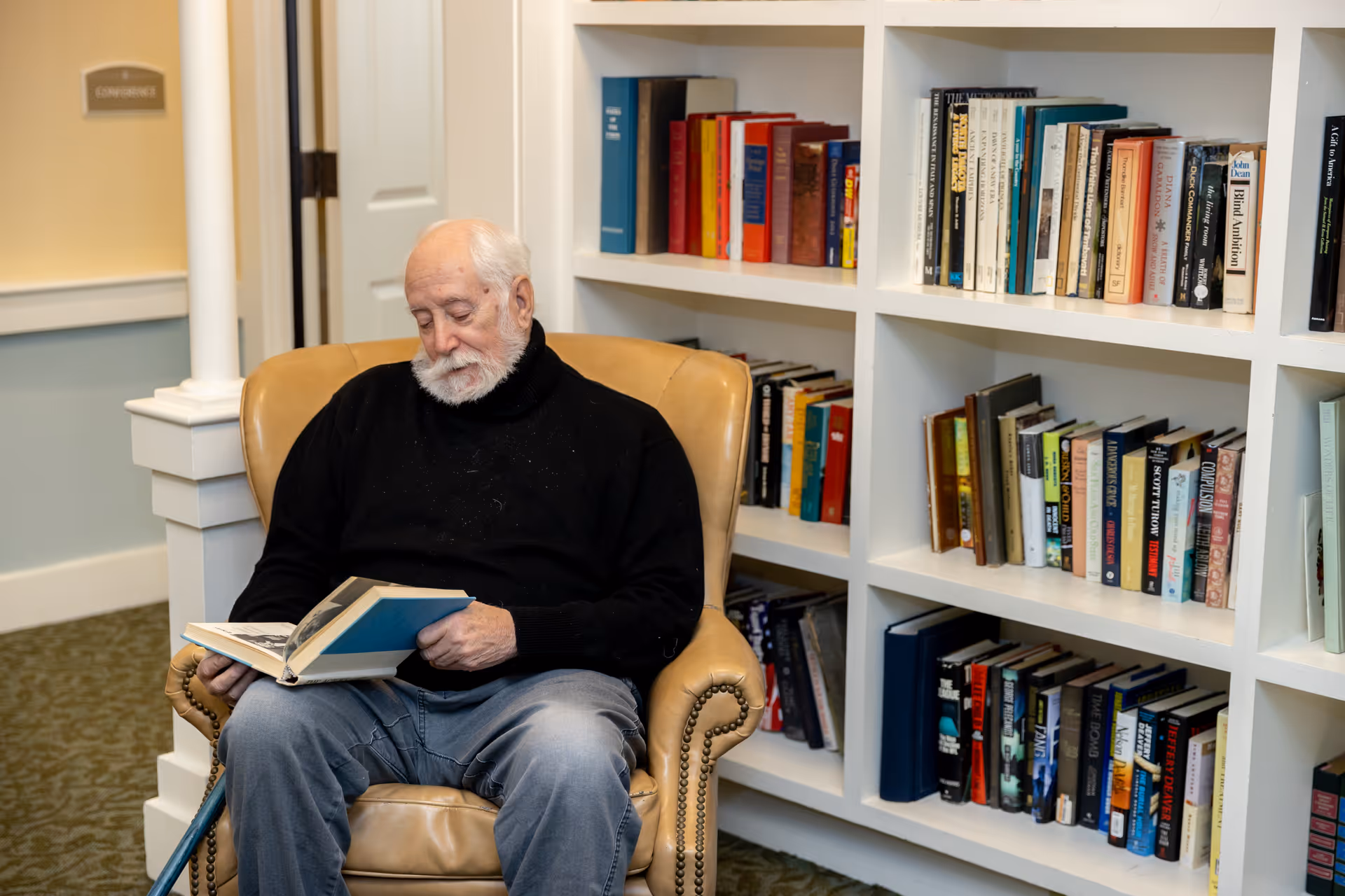 An elderly man reads a book while seated in a leather armchair next to built-in bookshelves in a communal interior space.