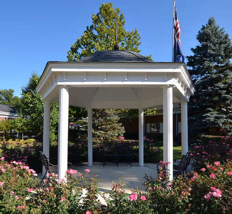 White hexagonal gazebo in a landscaped garden with pink roses, benches, and an American flag under a clear blue sky.