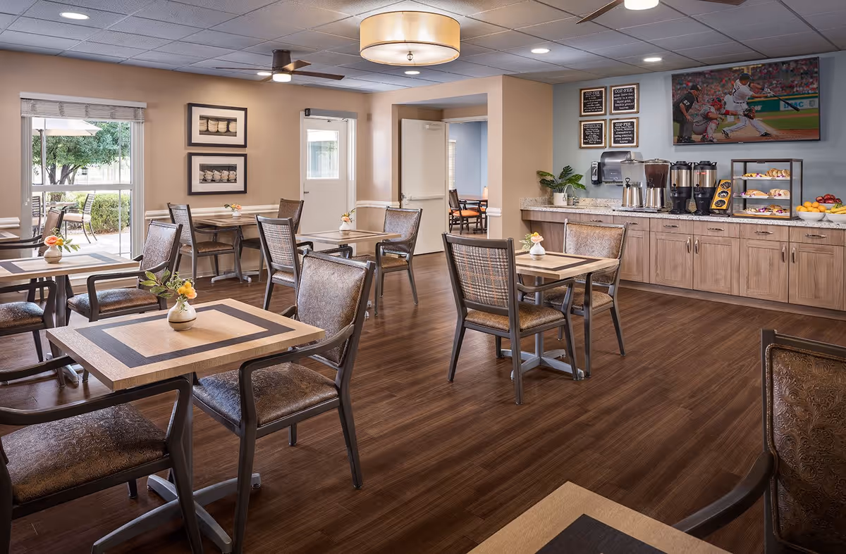 A bright and clean dining area in a senior living facility with several square tables and cushioned chairs arranged neatly. There is a counter along one wall with coffee dispensers, pastries, and fruit bowls. A flat-screen TV is mounted above the counter showing a baseball game. Large windows and a door provide natural light and a view of an outdoor patio with seating.