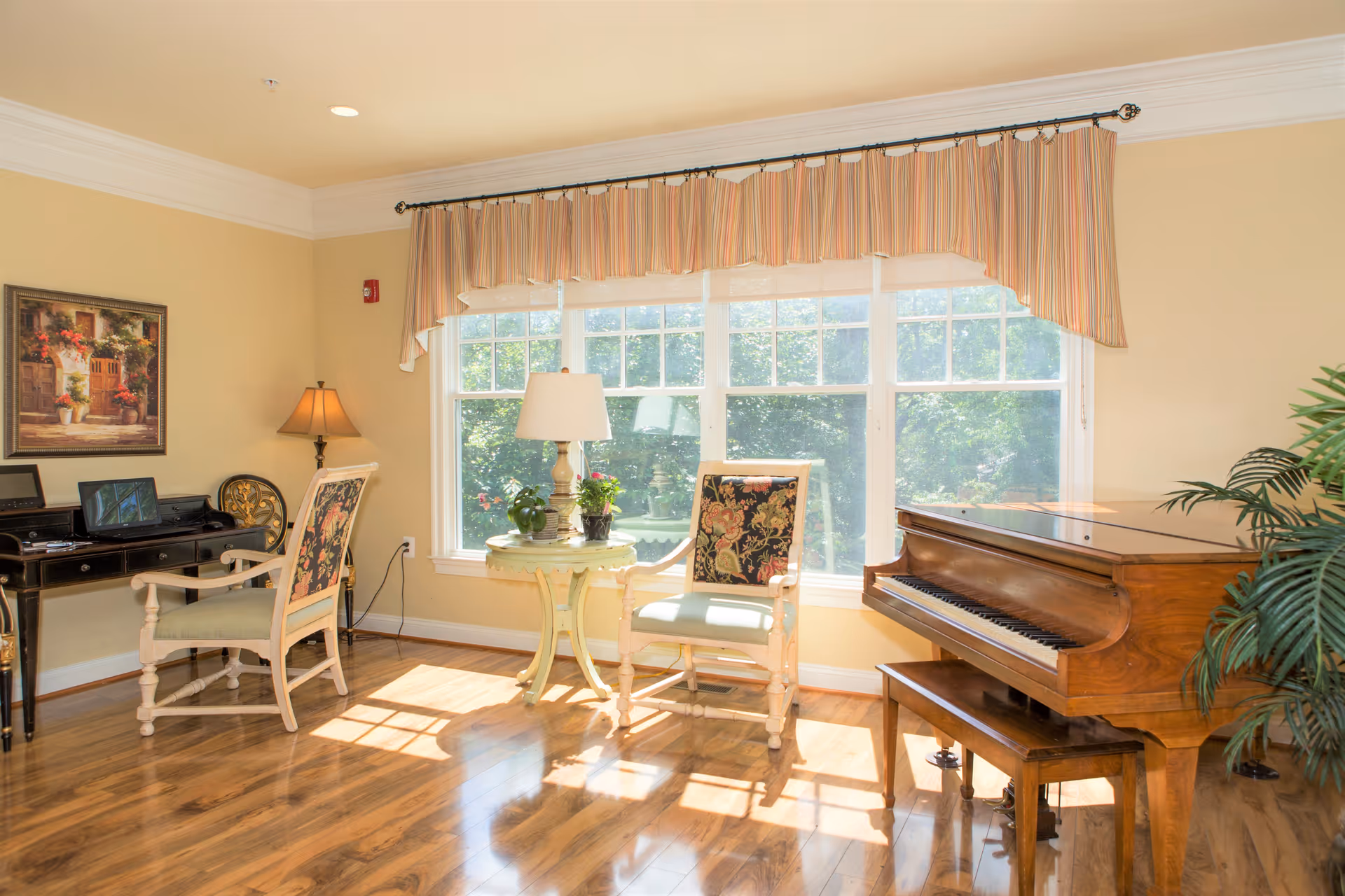 A bright and cozy living room with large windows letting in natural light. The room features a wooden floor, a grand piano with a matching bench, two upholstered chairs with floral patterns, a small round table with a lamp and potted plants, a desk with a computer, and a framed painting on the wall.