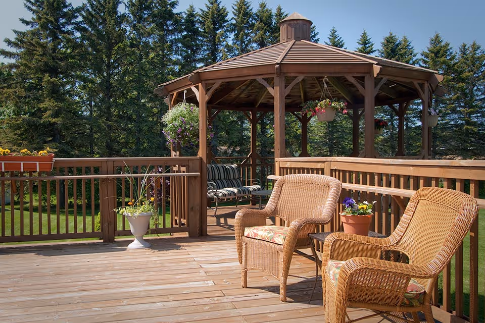 Outdoor wooden deck area with two wicker chairs and a small table with a potted plant. In the background, there is a wooden gazebo with hanging flower pots and a cushioned bench. Tall evergreen trees surround the area.