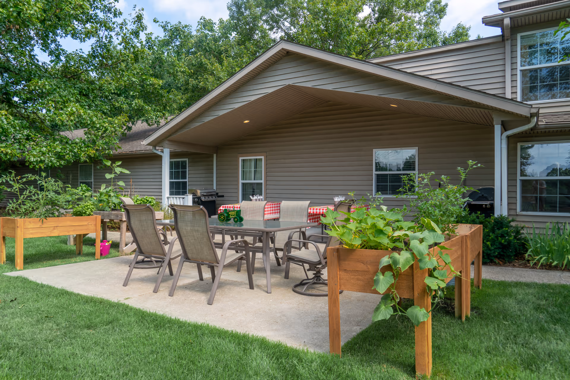 Outdoor patio area at Golden Orchards with a covered seating space featuring a glass table and six chairs. There are raised wooden garden beds with green plants on either side of the patio, and the building exterior is visible in the background with beige siding and windows.