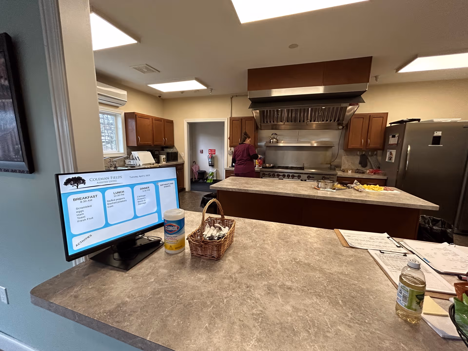 Interior view of a kitchen in Coleman Fields Assisted Living & Memory Care. A person in a maroon uniform is working near a large stove with an industrial exhaust hood. The kitchen has wooden cabinets, a refrigerator, and a countertop with various items including a basket, disinfectant wipes, a bottle of juice, and a monitor displaying the daily menu and activities.