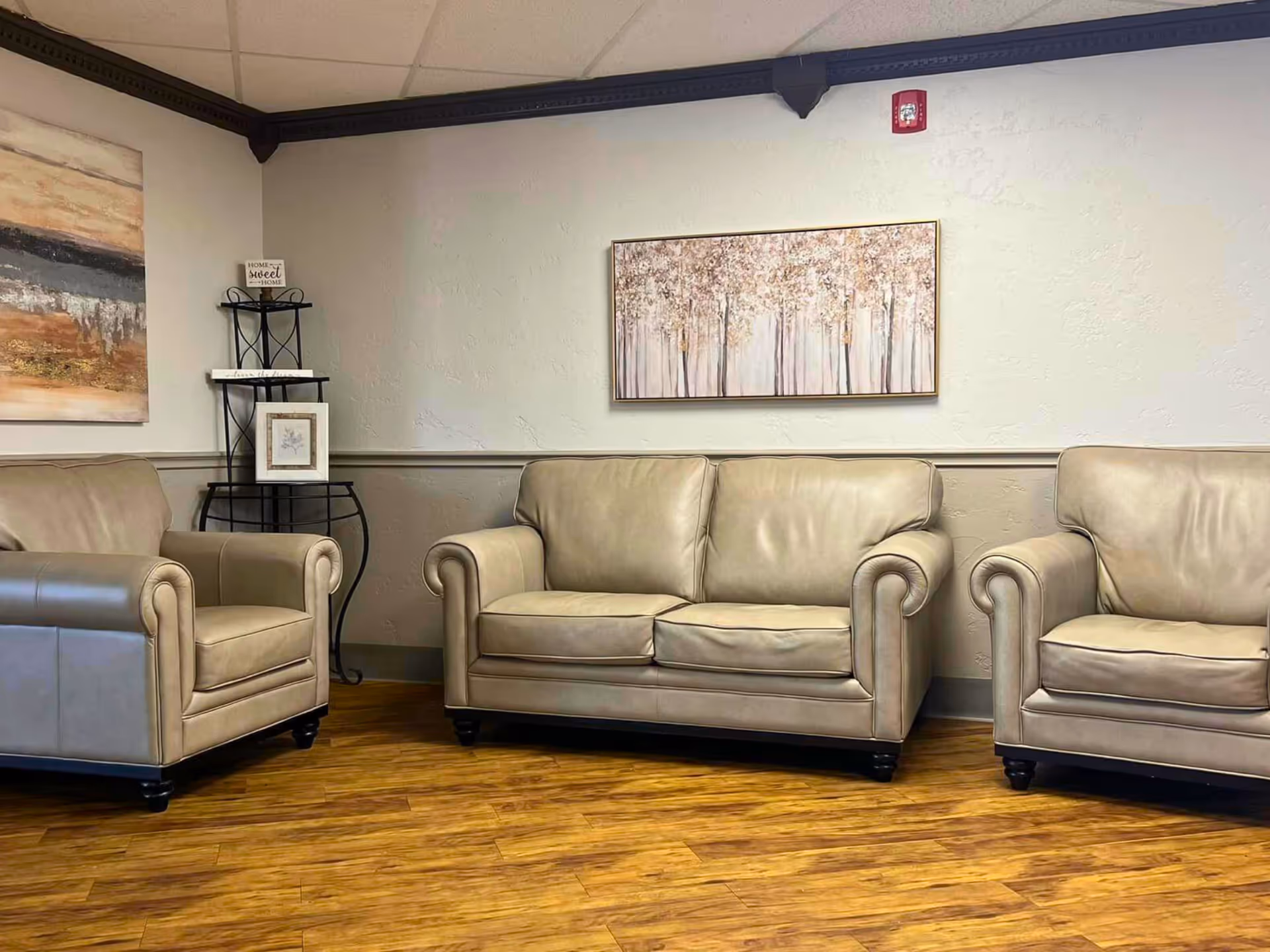 Seating area with three beige leather sofas, wood-look floor, and framed wall art in a nursing facility lounge.