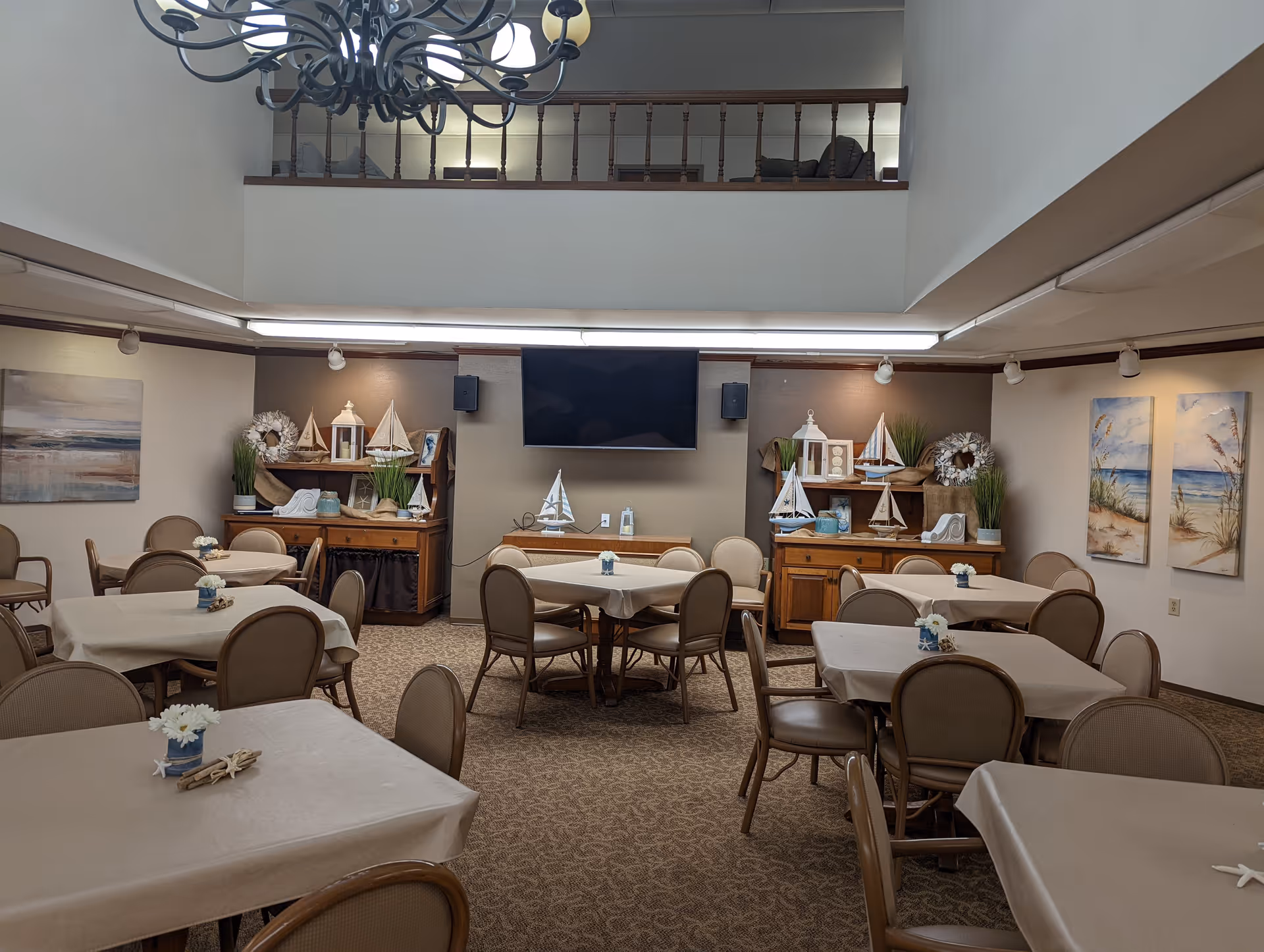Well-lit communal dining room with multiple tables and chairs, nautical decorations, and a wall-mounted TV beneath a mezzanine.