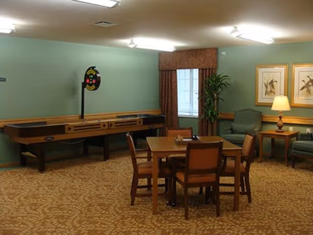 A common area in a senior living facility with a shuffleboard table against a green wall, a square wooden table with four chairs in the center, and a seating area with two armchairs, a side table with a lamp, and framed artwork on the wall. There is a window with curtains and a tall plant next to it.
