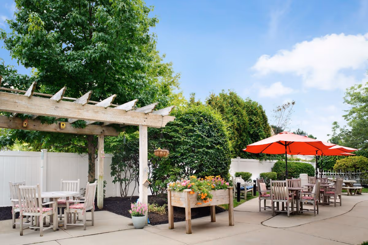 Outdoor courtyard with a wooden pergola, several dining tables and chairs, red patio umbrellas, and planters amid trees and shrubs.