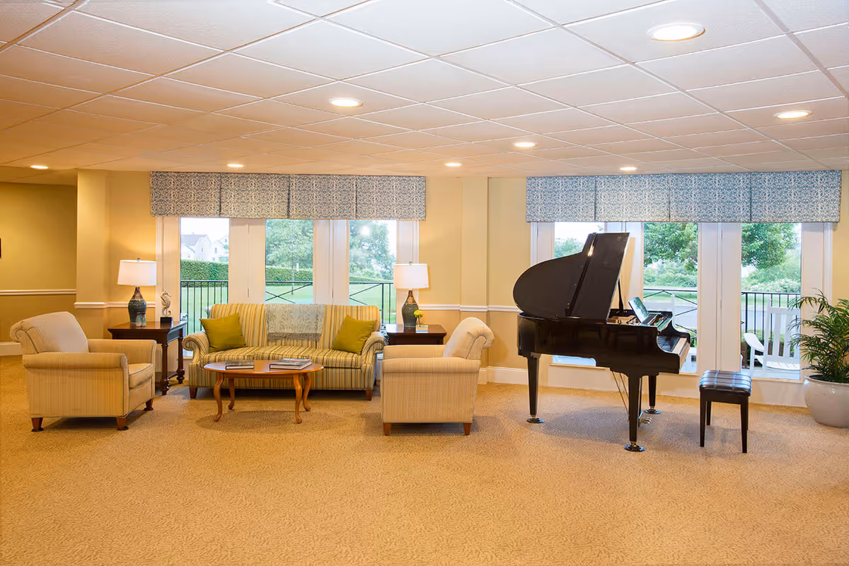 A bright and spacious living room area with beige carpet and cream-colored walls. The room features a striped sofa with green cushions, two beige armchairs, a wooden coffee table with books, two side tables with lamps, and a black grand piano with a matching bench. Large windows with patterned valances provide a view of greenery outside.