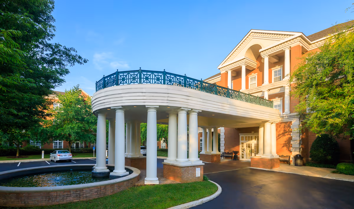 Exterior view of AdamsPlace senior living facility showing a grand entrance with white columns, a circular driveway, a small water feature, and a multi-story brick building with white trim and balconies under a clear blue sky.