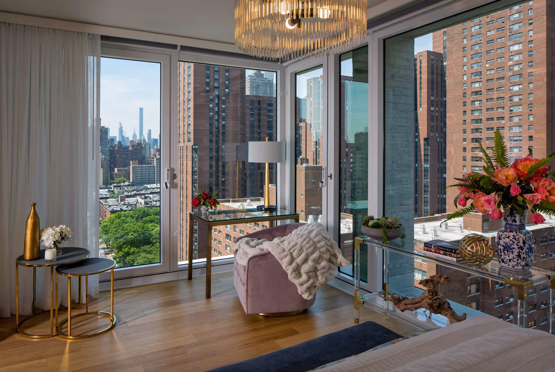 Sunlit corner sitting area of a bedroom with floor-to-ceiling windows overlooking the city, a pink armchair with a throw, side tables, and floral decor.