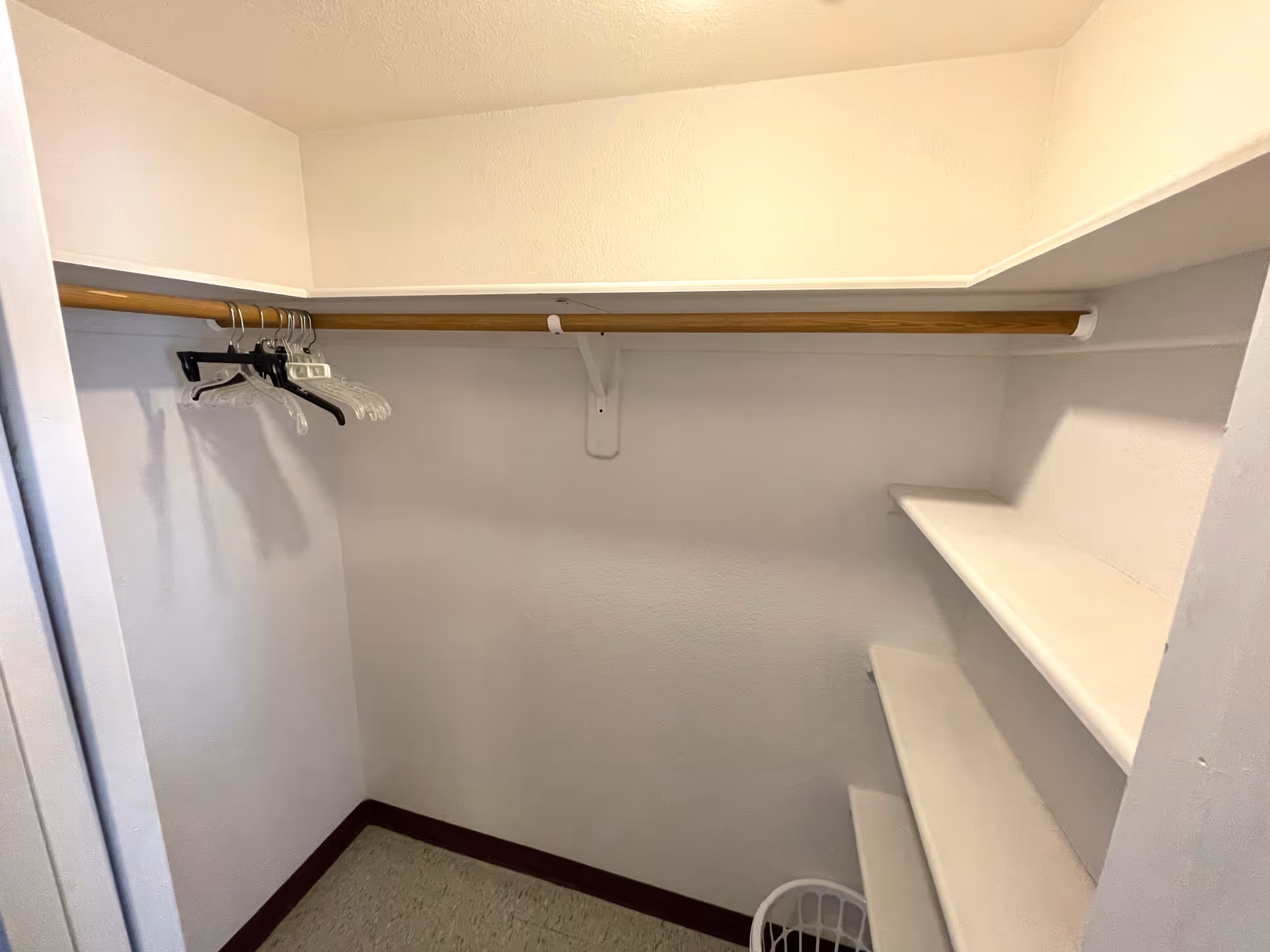 Empty walk-in closet with a wooden hanging rod holding several hangers on the left side, white shelves on the right side, and a white laundry basket on the floor.