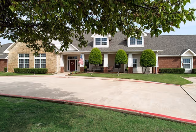 Front exterior of a single-story senior living building with a covered porch, American flag, trimmed shrubs, and a curved driveway.