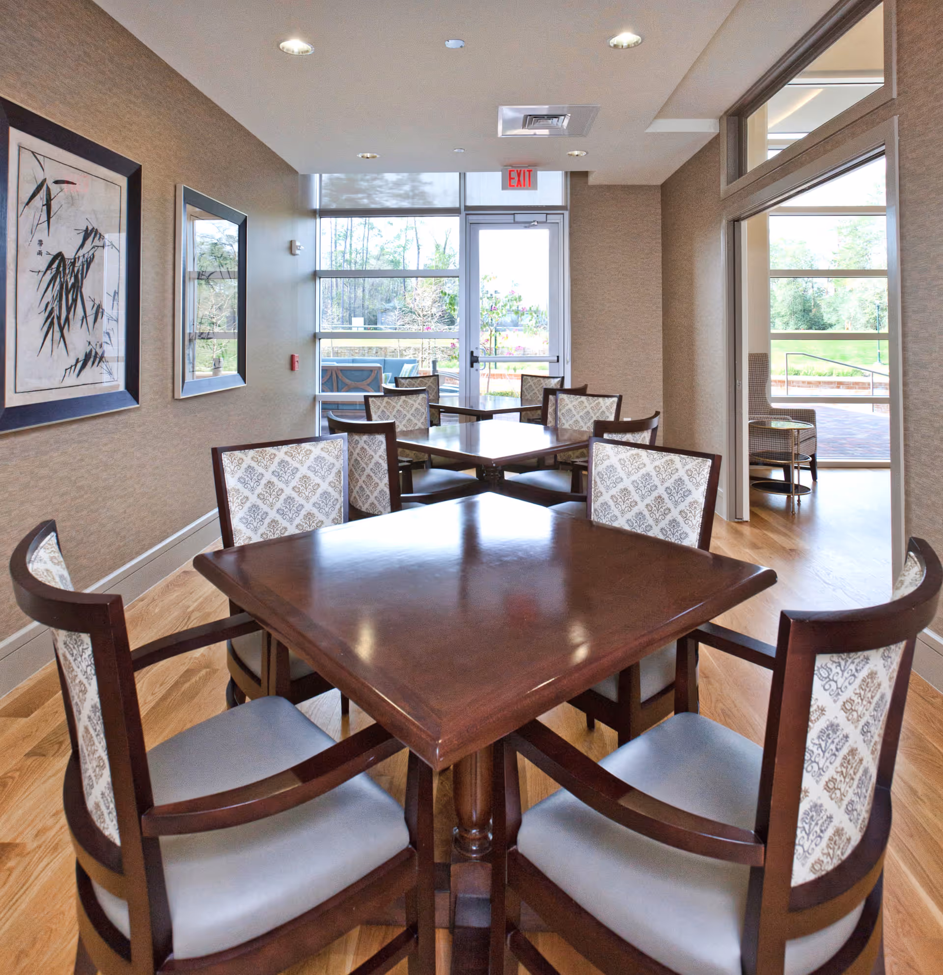 Interior view of a dining area with wooden tables and cushioned chairs featuring patterned backs. The room has light-colored walls with framed artwork, large windows, and a glass door leading outside. Natural light brightens the space, and there is a glimpse of an outdoor patio area through the windows.