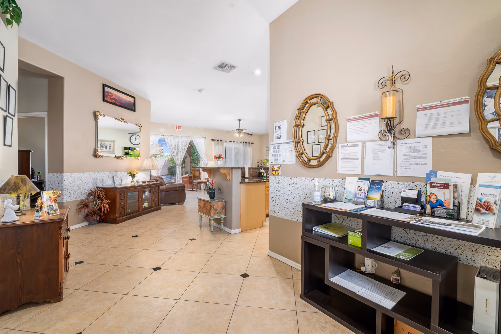 Interior view of a senior living facility showing a spacious common area with tiled floors, beige walls, and various wooden furniture pieces including cabinets and shelves. The area features decorative mirrors, informational brochures, wall-mounted documents, a lamp, and a small table with a plant. In the background, there is a seating area with a sofa and large windows with sheer curtains allowing natural light to enter.