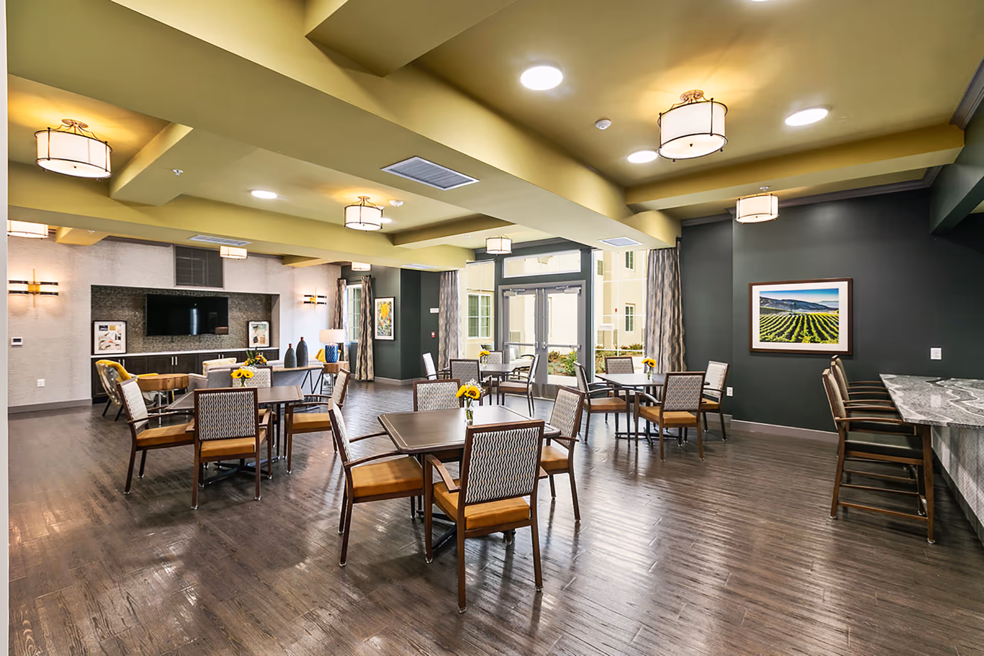 Spacious communal dining and lounge area with multiple tables and chairs, a TV wall, bar seating, and large windows.