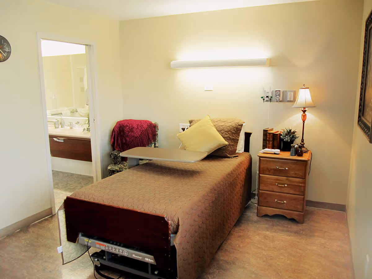 A furnished single-occupancy bedroom with a hospital-style bed, overbed table, nightstand with lamp, and an open doorway leading to a bathroom.