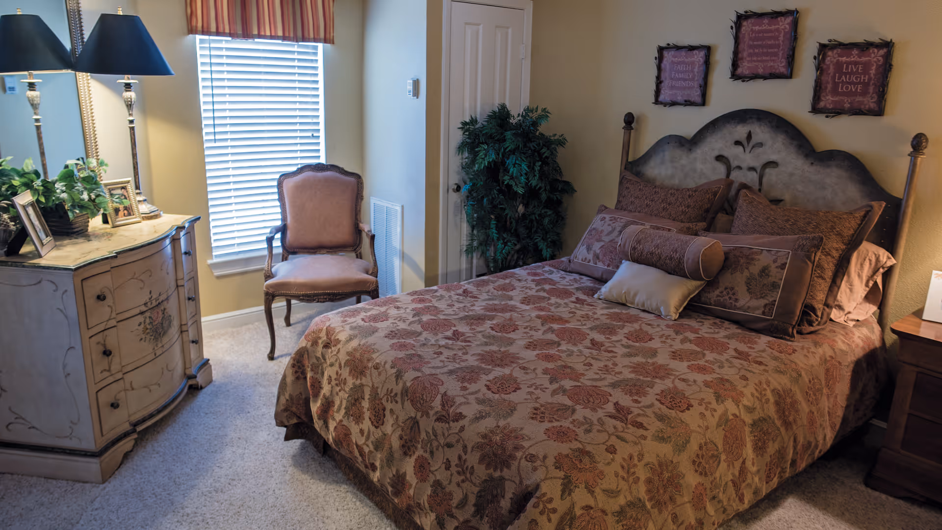 A cozy bedroom with a floral patterned bedspread on a bed with multiple pillows. There is a vintage-style armchair near a window with blinds and a striped valance. A decorative dresser with two lamps and framed photos is on the left side. On the wall above the bed are three framed inspirational quotes. A green potted plant is placed near a closed door.