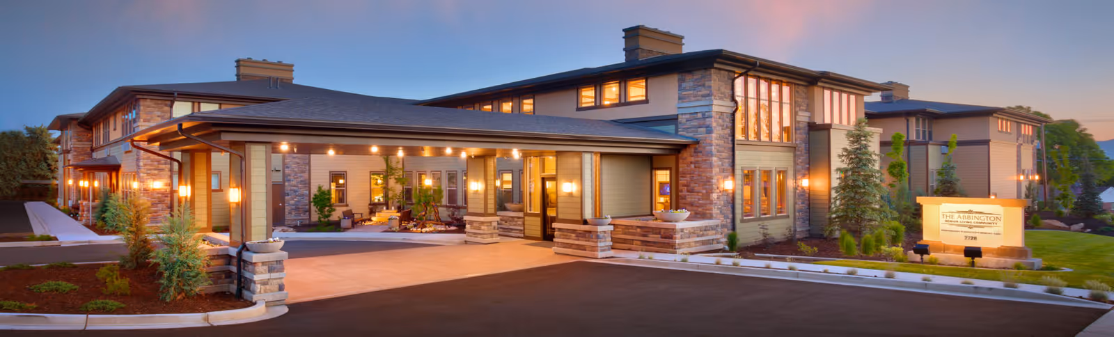 Exterior view of The Abbington at Holladay senior living community building during dusk, showing a well-lit entrance with a covered driveway, landscaped greenery, and a sign with the facility's name.