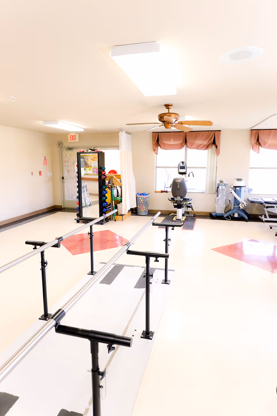 A bright rehabilitation room with parallel bars for walking exercises, exercise equipment including a stationary bike, weights, and therapy machines. The room has large windows with brown valances, a ceiling fan, and fluorescent lighting.