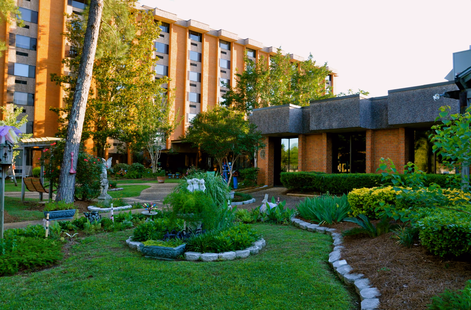 A landscaped garden area with green grass, various plants, shrubs, and small statues in front of a multi-story brick building with large windows. Trees and a paved walkway are also visible in the garden.
