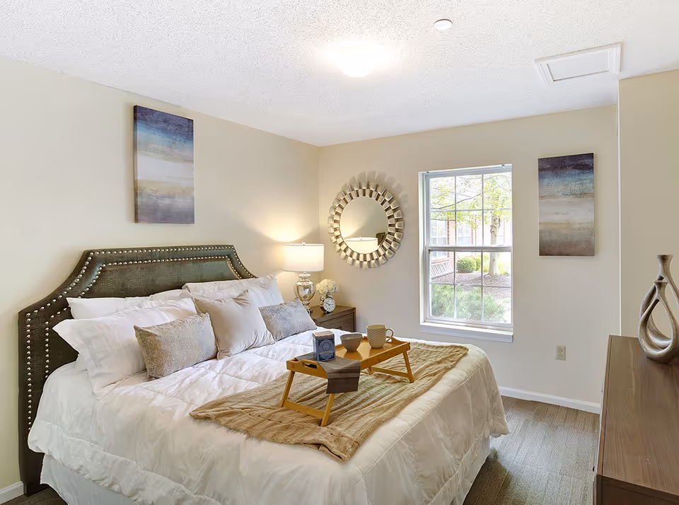 A bright decorated bedroom with a neatly made bed, throw pillows and a breakfast tray, a round wall mirror, nightstand with a lamp, and a window.