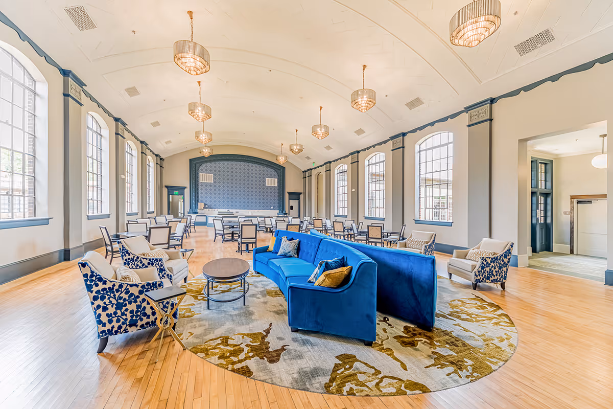 Spacious senior living common area with large arched windows, wooden floors, and a high vaulted ceiling with multiple chandeliers. The room features a blue velvet sectional sofa, patterned armchairs, a round rug, and several dining tables and chairs arranged towards the back.