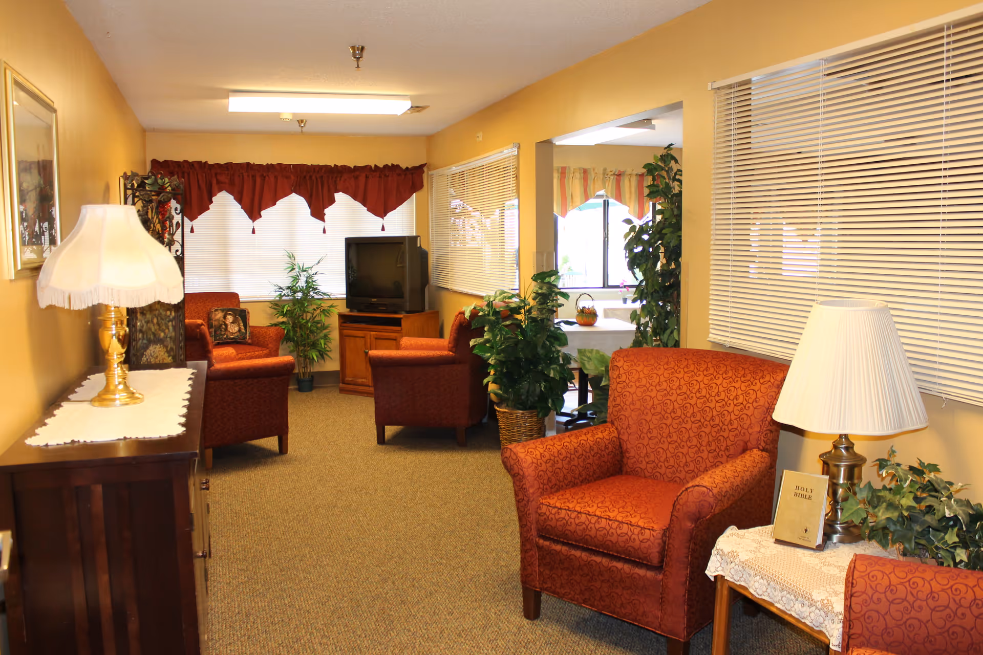 A cozy living room area with beige walls and carpeted floor. The room contains several red patterned armchairs, a wooden TV stand with an older-style television, and various green plants. There are two table lamps with white shades, one on a wooden side table with a Holy Bible and some greenery, and another on a wooden cabinet with a lace runner. The windows have white blinds and red valances, and there is an open doorway leading to a dining area with a table and chairs.