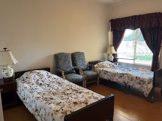 Shared bedroom with two twin beds covered in floral bedspreads, two upholstered chairs between them, bedside tables with lamps, and a curtained window.