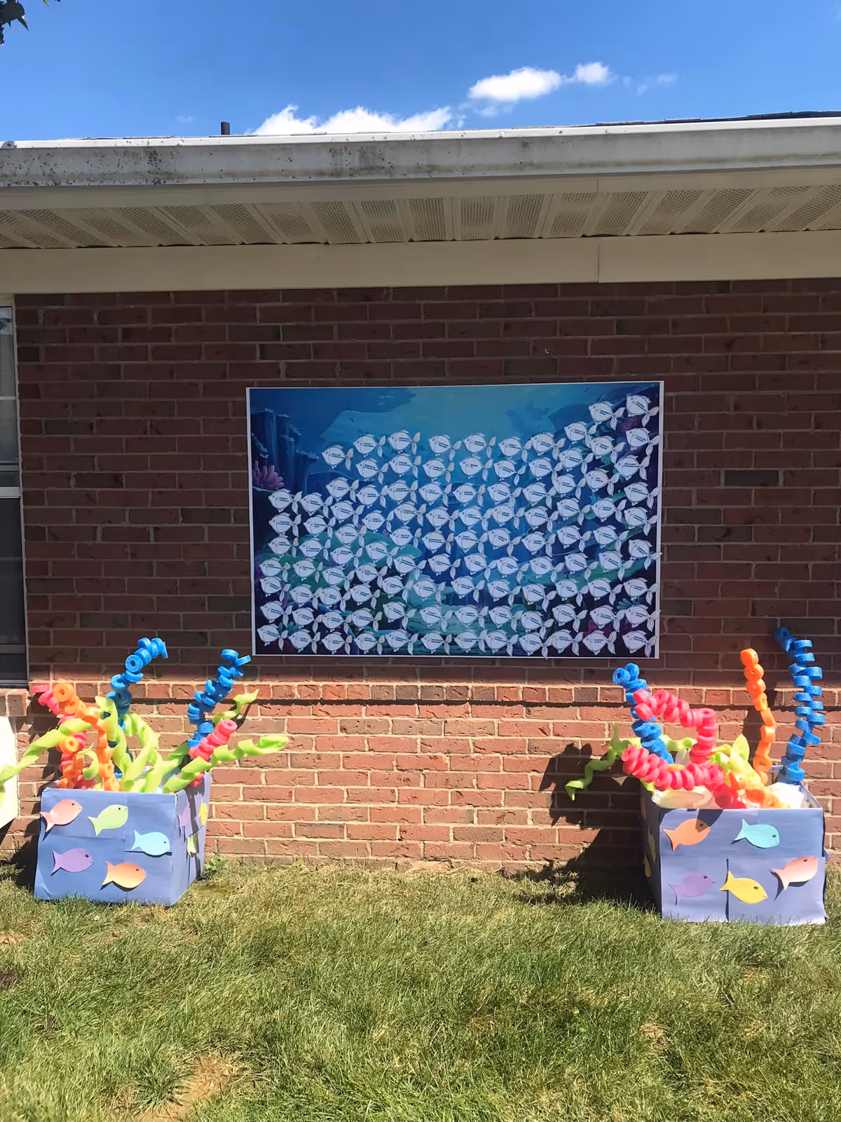 Outdoor scene showing a brick wall with a large poster depicting a school of fish. Below the poster, there are two decorative boxes on the grass, each filled with colorful pool noodles arranged to resemble underwater plants. The sky is clear with a few clouds visible above the roofline.