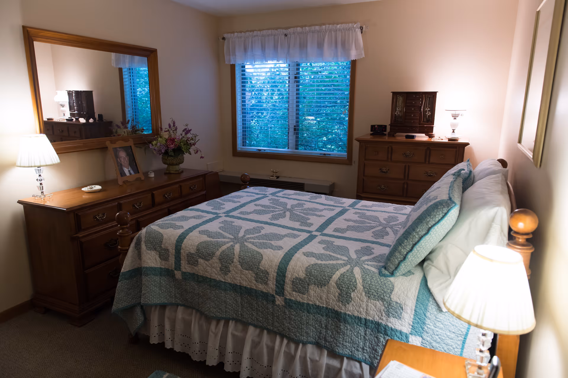 A cozy bedroom with a wooden bed frame and a quilted bedspread in white and teal patterns. There are two wooden dressers, one with a large mirror above it, a framed photo, a floral arrangement, and a lamp. The other dresser has a small jewelry cabinet and a lamp. A window with blinds and a white valance lets in natural light, and the walls are painted a soft beige color.