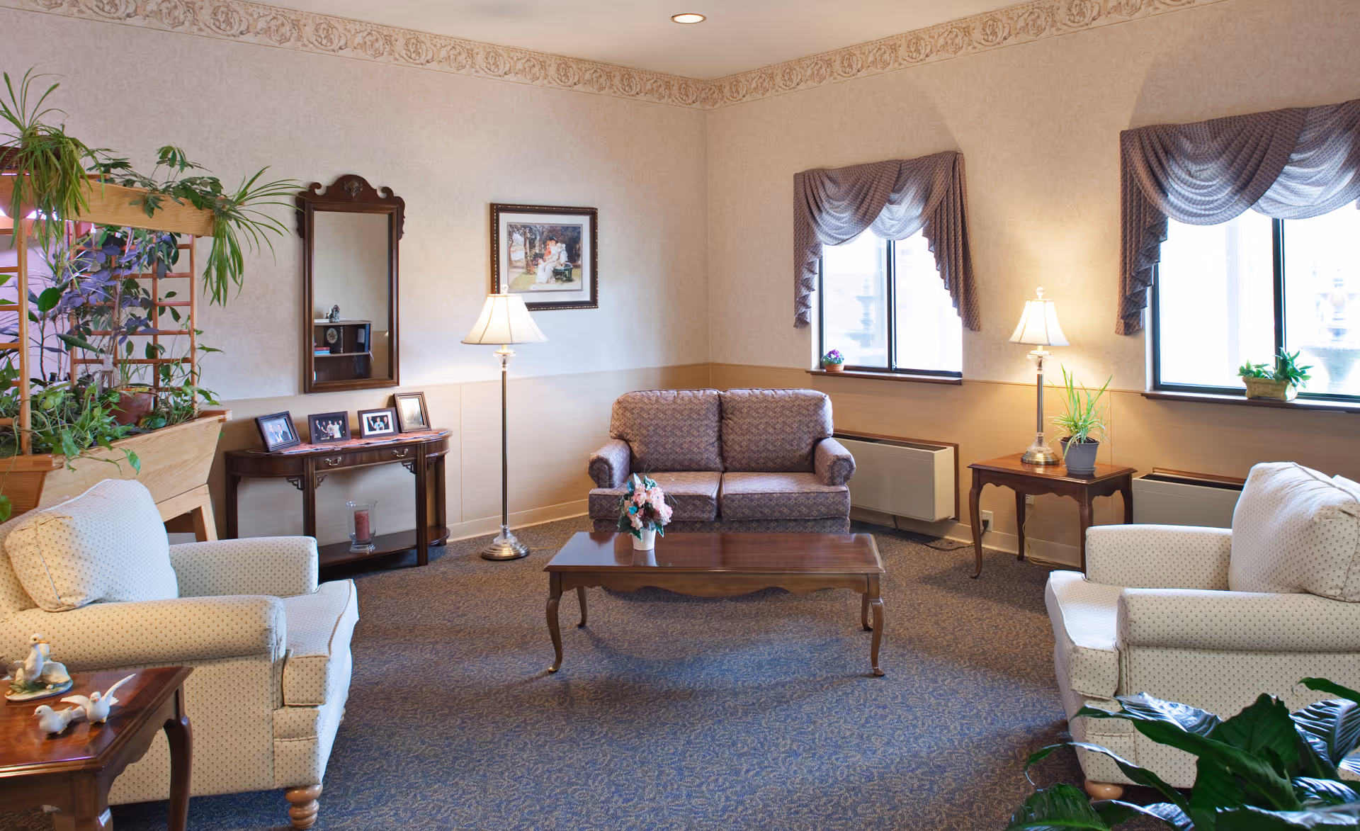 A cozy living room with two white armchairs and a patterned loveseat arranged around a wooden coffee table with a flower vase. The room has two windows with purple valances, two floor lamps, a wooden side table with framed photos, a mirror, and several green plants. The walls are light-colored with a decorative border near the ceiling.
