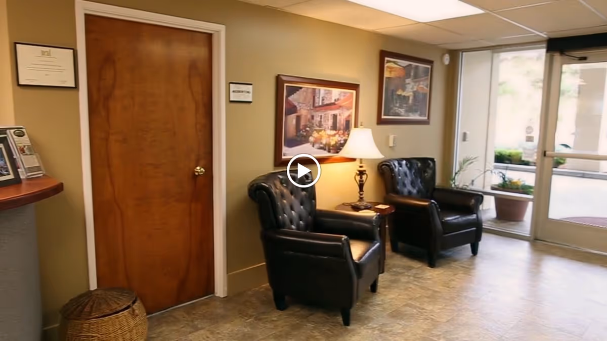 A small waiting area with two black leather armchairs separated by a wooden side table with a lamp. There are framed pictures on the beige walls, a wooden door, and a glass door leading outside with potted plants visible through the glass.