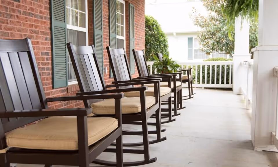 A row of cushioned wooden rocking chairs on a covered front porch with a brick wall and white railing.