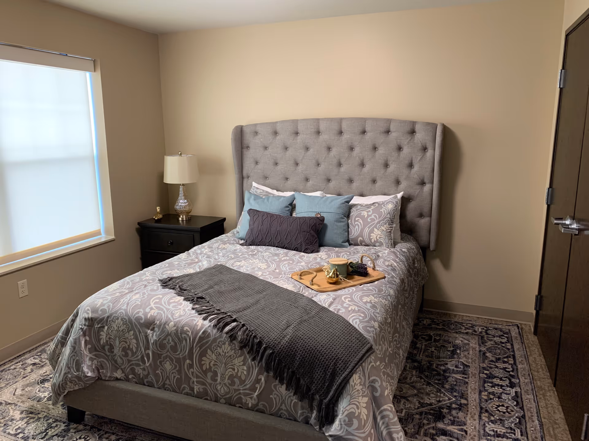 A neatly made bed with a tufted gray headboard in a bedroom. The bed has patterned bedding with multiple pillows in shades of blue, gray, and white, and a dark gray throw blanket at the foot. A wooden tray with a small teapot, cup, and lavender is placed on the bed. To the left of the bed is a black nightstand with a decorative lamp and a small golden bird figurine. A window with a closed white blind is on the left wall, and a closed wooden door is on the right. The floor has a patterned area rug.