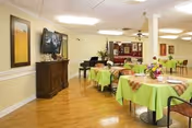 Interior view of a dining room in a senior living facility with round tables covered in green tablecloths and plaid runners, each decorated with flower centerpieces. There are chairs around the tables, a wooden cabinet with a flat-screen TV mounted above it, and a piano in the background. The room has wooden flooring, light-colored walls, and ceiling lights.