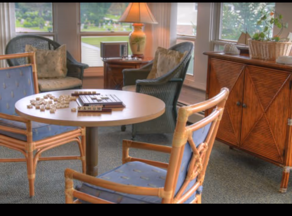 Sunlit sitting area with wicker and rattan chairs around a round table holding a word game and tiles, beside a lamp and wooden cabinet by windows.