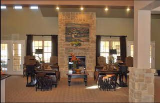 Spacious common room with a central stone fireplace, seating groups, side tables and lamps, and a painting above the mantel.