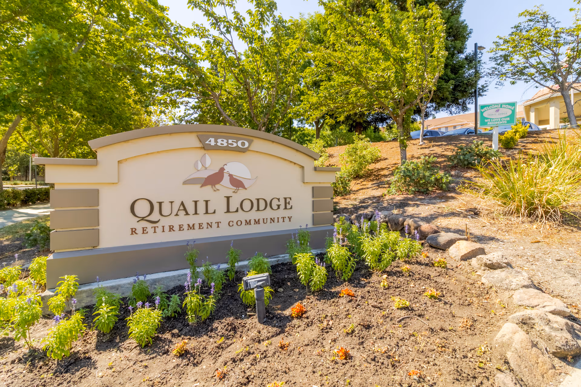 Outdoor view of the entrance sign for Quail Lodge Retirement Community with the address number 4850. The sign is surrounded by a landscaped garden with small plants and flowers, and there are trees and a parking area in the background under a clear blue sky.