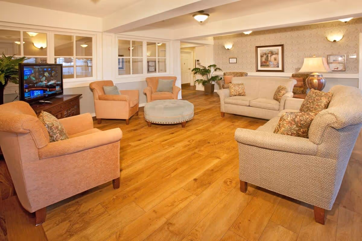 A cozy living room area with wooden flooring, featuring two beige sofas with patterned cushions, two peach armchairs with blue cushions, a round blue ottoman in the center, a wooden TV stand with a television, potted plants, and framed artwork on the wallpapered walls.