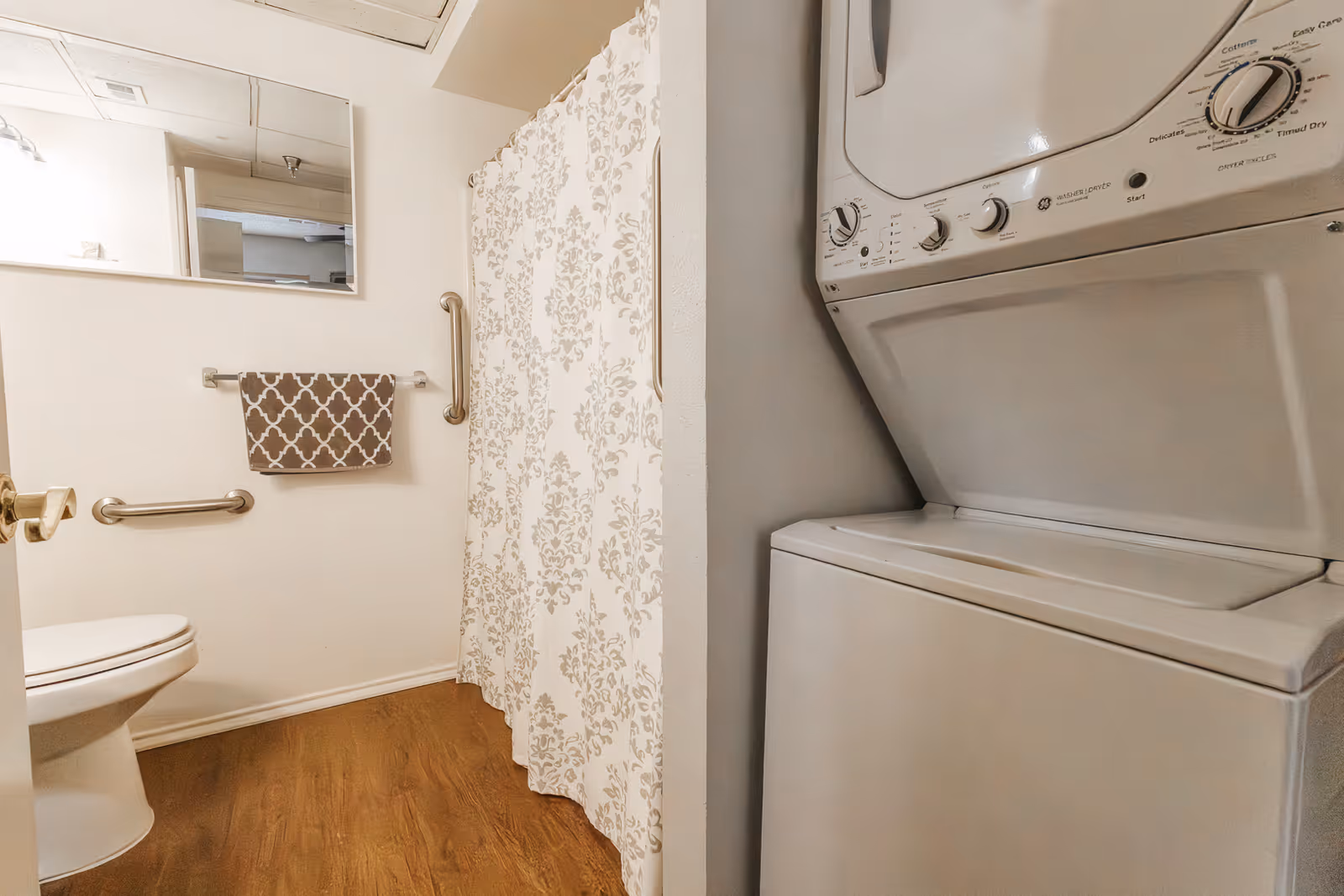 A bathroom with a toilet, a towel hanging on a rack, a large mirror above the towel, a shower with a patterned curtain, and a stacked washer and dryer unit on the right side.