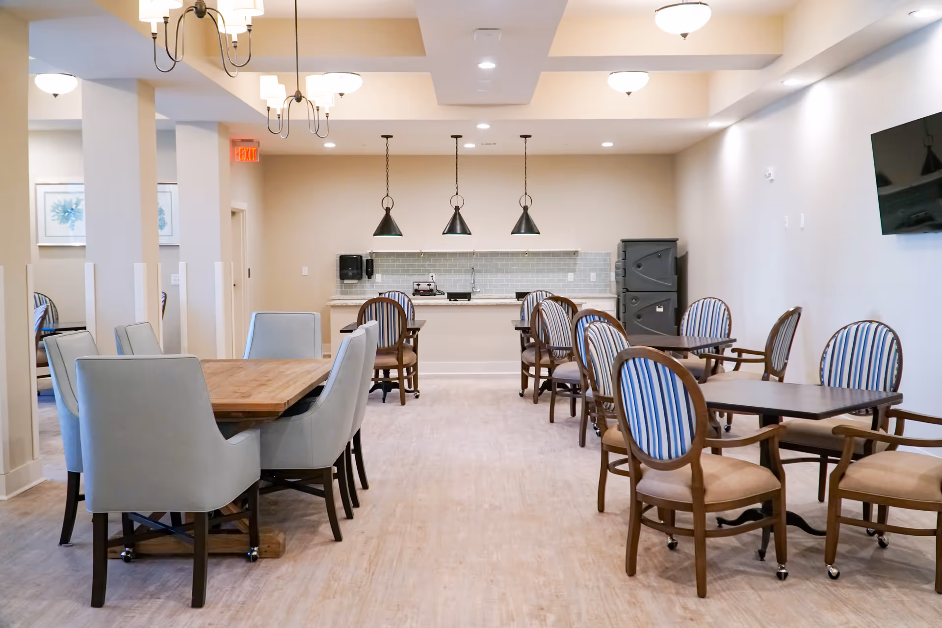 A bright dining room with multiple tables and chairs. There are two types of chairs: cushioned light blue chairs around a wooden table on the left, and wooden chairs with striped upholstery around smaller dark wood tables on the right. The back wall features a counter with a light blue tiled backsplash, three hanging pendant lights, and a black cabinet. The ceiling has recessed lighting and chandeliers, and a flat-screen TV is mounted on the right wall.