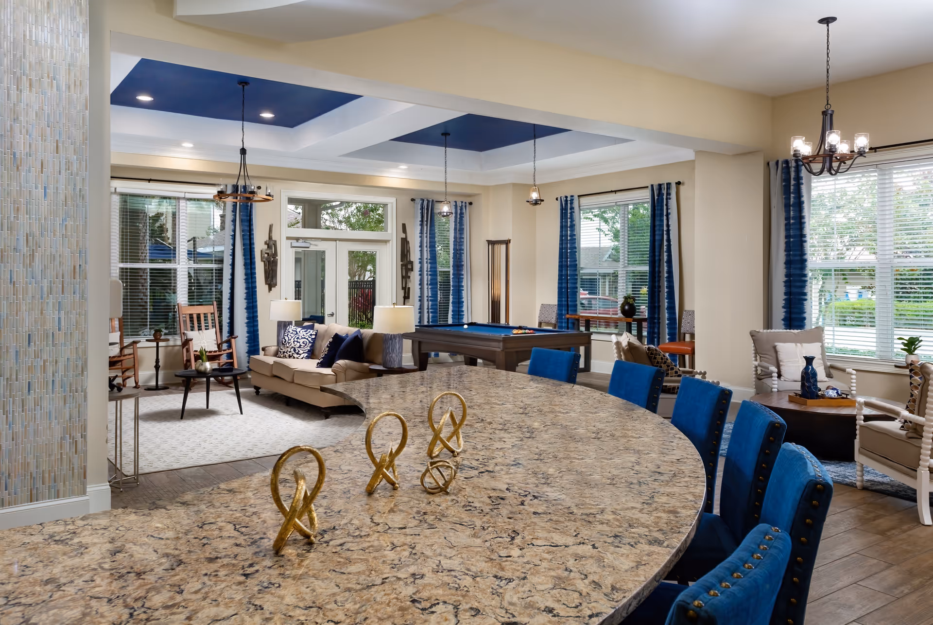 A spacious and well-lit common area in Album Monterey Pointe featuring a large granite countertop with decorative golden sculptures, blue upholstered chairs, a beige sofa with blue and white pillows, a pool table, rocking chairs, and large windows with blue curtains allowing natural light to fill the room.