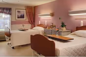 A hospital-style room with two single beds, each with white bedding and wooden headboards. There is a window with curtains on the left side, a framed picture on the wall, and a small table with flowers between the beds. The walls are painted a soft pink color, and there are wall-mounted lights above each bed.