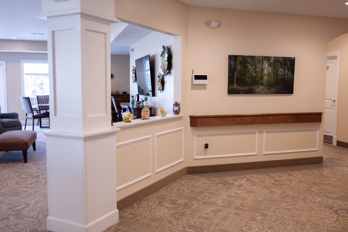 Interior view of a senior living facility showing a hallway with beige walls and patterned carpet. There is a white column in the foreground, a wooden handrail along the wall, a thermostat, and a framed picture of a forest on the wall. In the background, there is a seating area with chairs and a TV mounted on the wall.