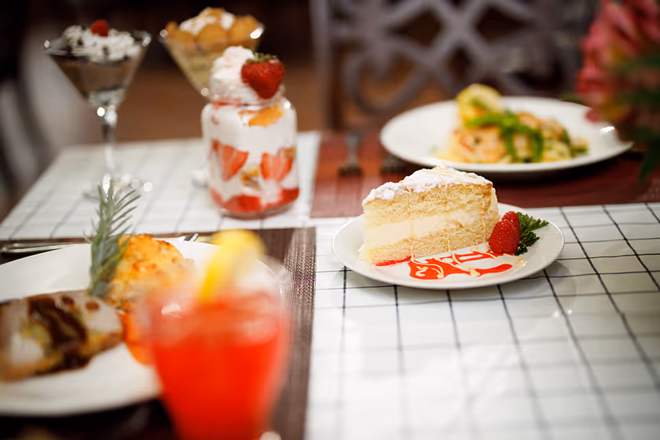 A table set with various plated desserts and dishes, including a slice of layered cake with strawberry garnish and sauce, a dessert in a glass jar with strawberries and whipped cream, and other plated food items blurred in the background.