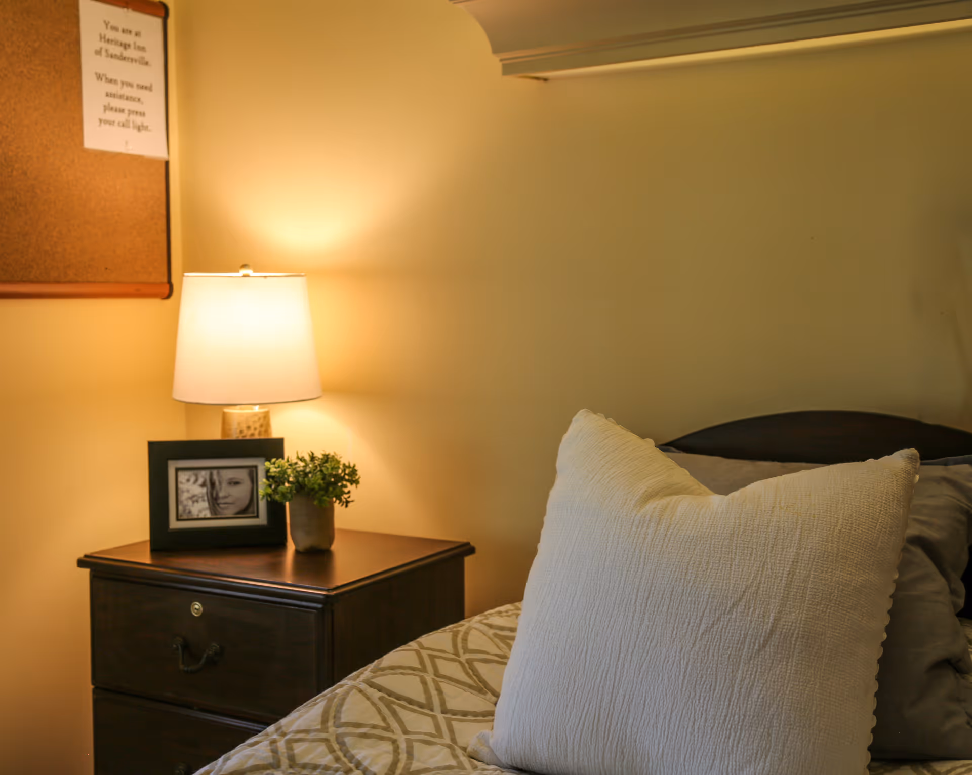 A cozy bedroom corner with a wooden nightstand holding a lit table lamp, a small potted plant, and a framed black and white photo. The bed next to the nightstand has a large white pillow and patterned bedding. A corkboard with a notice is partially visible on the wall above the nightstand.